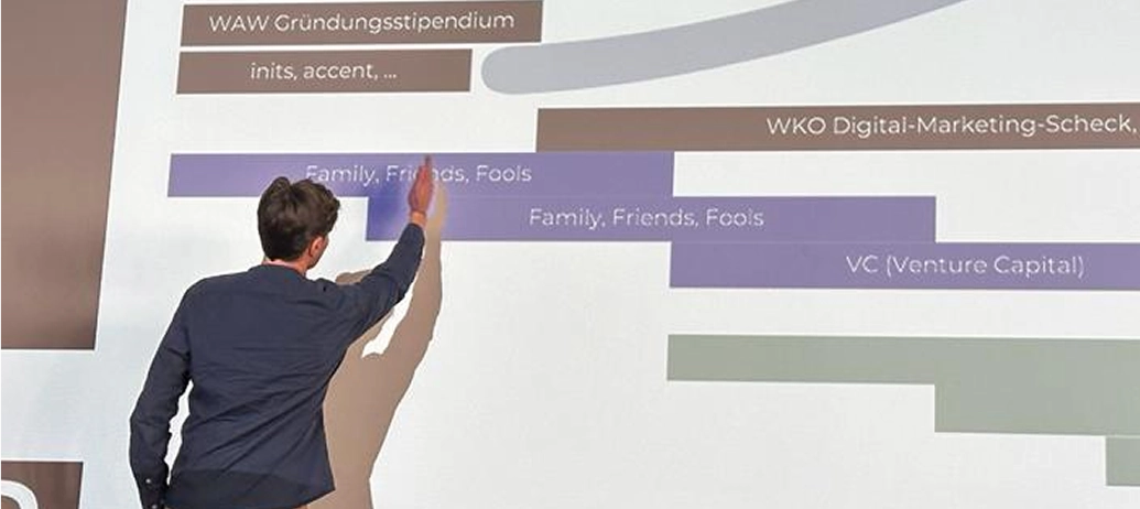 Manuel Rainer, Managing Partner und Förderberater bei MINTED, hält einen Vortrag über Förderungen auf der Bühne und zeigt auf den Text "Family, Friends, Fools" in seiner Präsentation